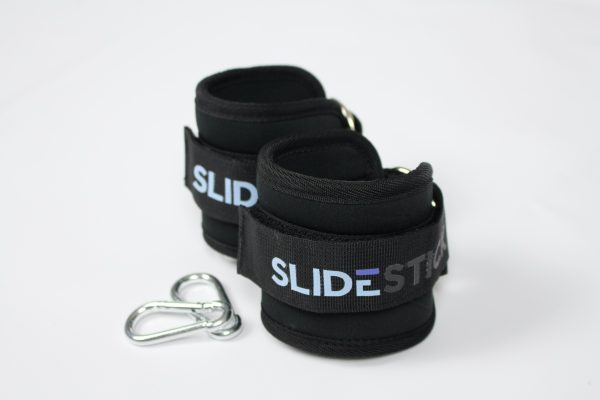 Zwei schwarze Fußschlaufen mit dem SlideStick-Logo, die hintereinander aufgestellt sind. Links daneben liegen zwei Karabinerhaken
